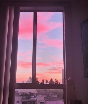 Ikkunasta avautuu näkymä auringonlaskun värittämälle taivaalle ja sen alla oleviin rakennuksiin.