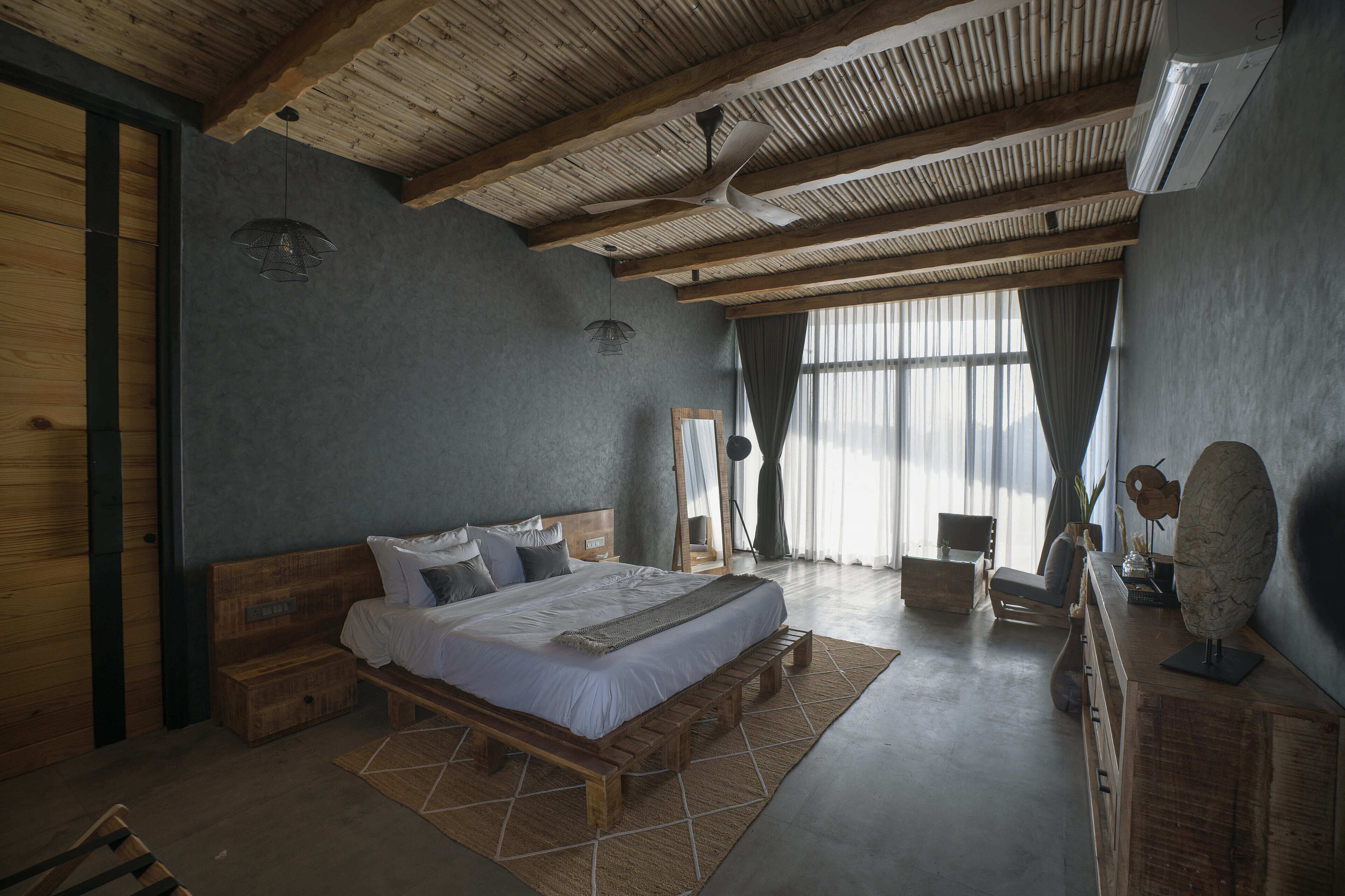 A spacious bedroom with a large bed, wooden headboard, and a wooden bench. There is a desk with a mirror and a decorative object on it. The room has a high ceiling with wooden beams and an air conditioning unit.