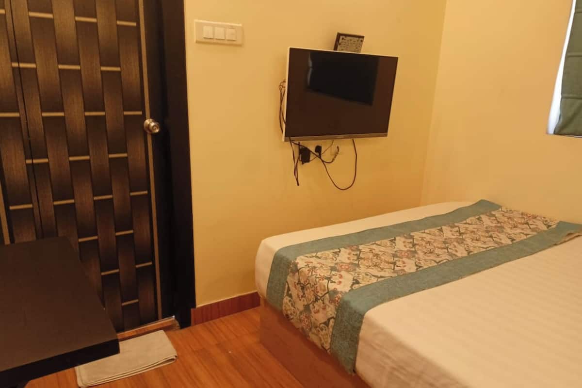 Una habitación con puerta de madera, cama con colcha estampada, televisor montado en la pared y una mesita con mantel blanco.