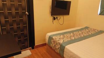 Una habitación con puerta de madera, cama con colcha estampada, televisor montado en la pared y una mesita con mantel blanco.