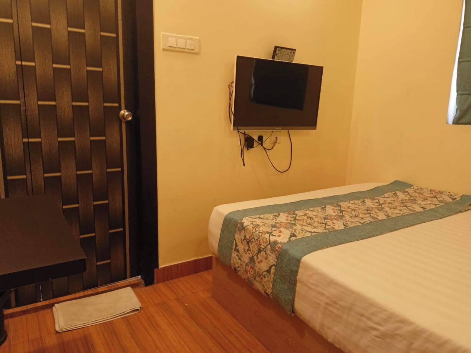 Una habitación con puerta de madera, una cama con cubrecama floral, un escritorio con silla y un televisor montado.