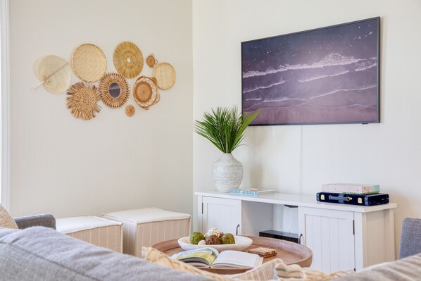 Una sala de estar con un sofá blanco, una mesa de centro con un libro y objetos decorativos, un televisor montado en la pared y un estante con una planta y una maleta.
