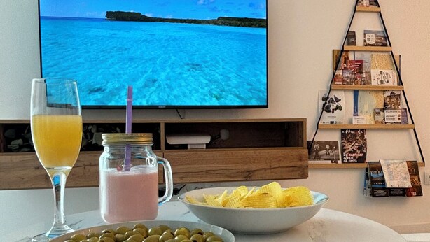 Ein Esstisch mit Oliven, Chips und einem Getränk, ein Fernseher zeigt eine Strandansicht und ein Bücherregal mit Büchern und Zeitschriften.