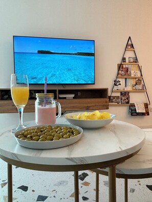 Una mesa de comedor con un tazón de aceitunas, patatas fritas y una bebida, un televisor que muestra una escena de playa y una estantería con libros y revistas.