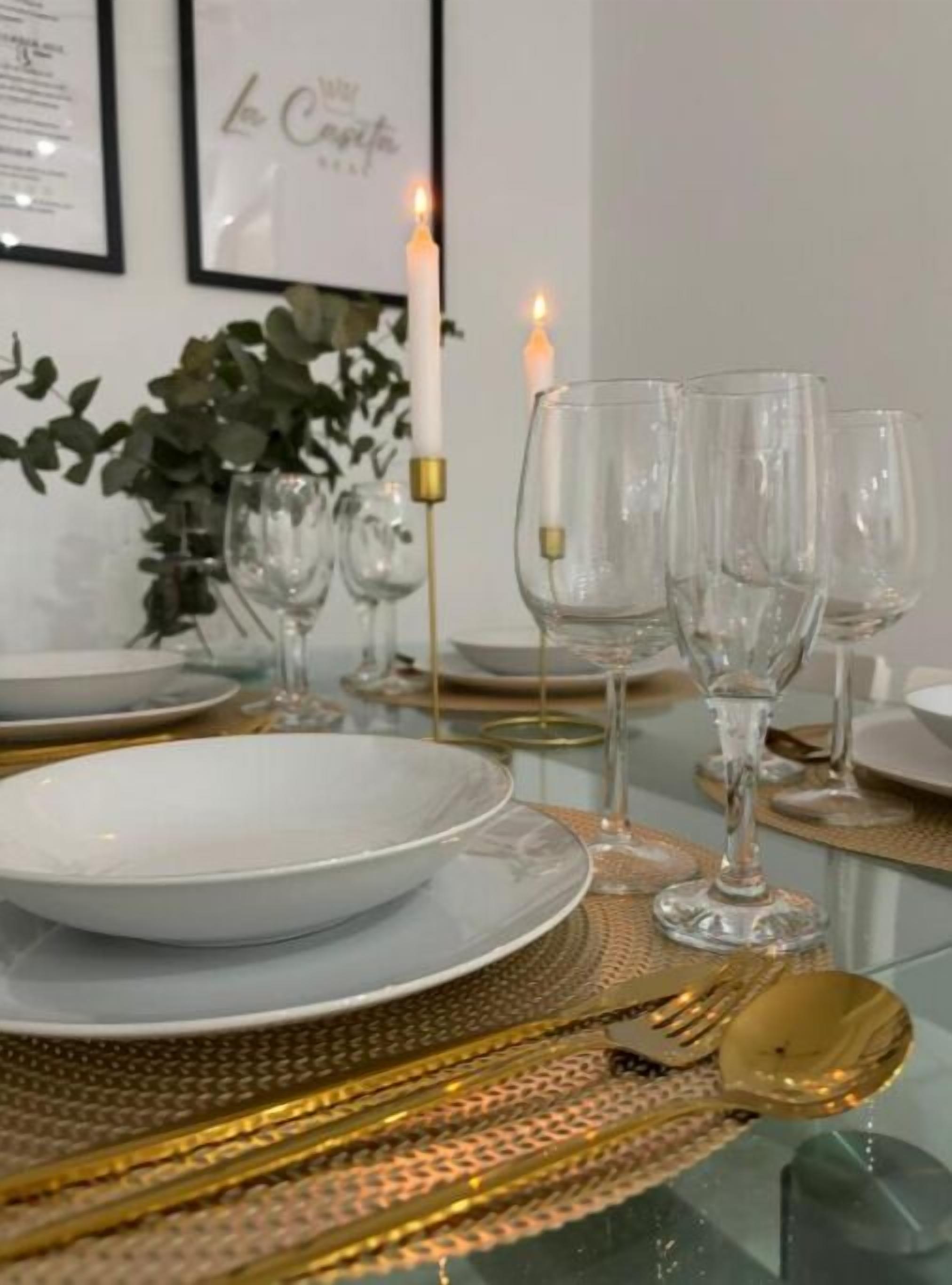 Una mesa de comedor con platos blancos, copas de vino transparentes y cubiertos dorados.