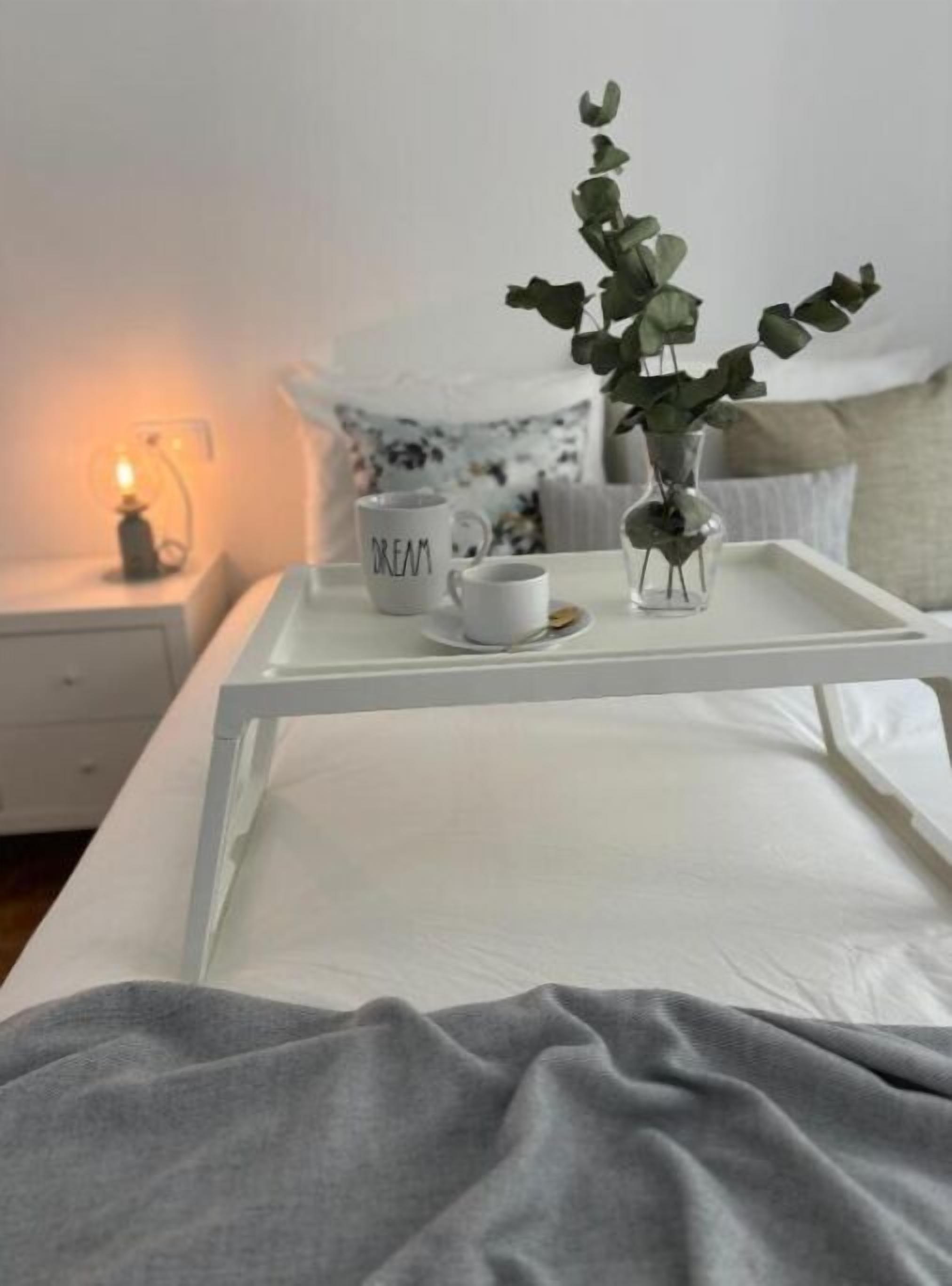 En snyggt bäddad säng med en vit madrass, en grå filt och ett vitt nattduksbord med en liten lampa, en mugg med ordet ”DRÖM” på och en vas med gröna blad.