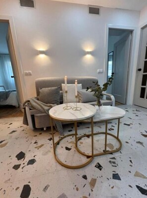 Un salon moderne avec un canapé gris, deux petites tables rondes blanches à cadre doré et un sol en terrazzo.
