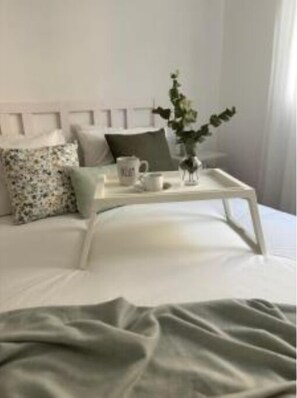 Un lit bien fait, avec une tête de lit blanche, un plaid vert et un plateau sur lequel se trouvent une tasse et une petite plante.
