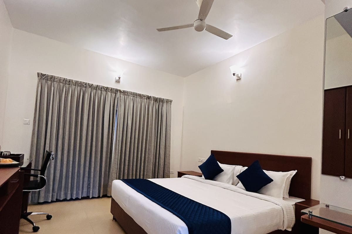 A hotel room with a bed, a desk, a chair, a ceiling fan, and a large window with curtains.