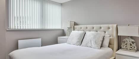 A bedroom with a bed, two bedside tables, a nightstand, a window with blinds, and a wall-mounted lamp.