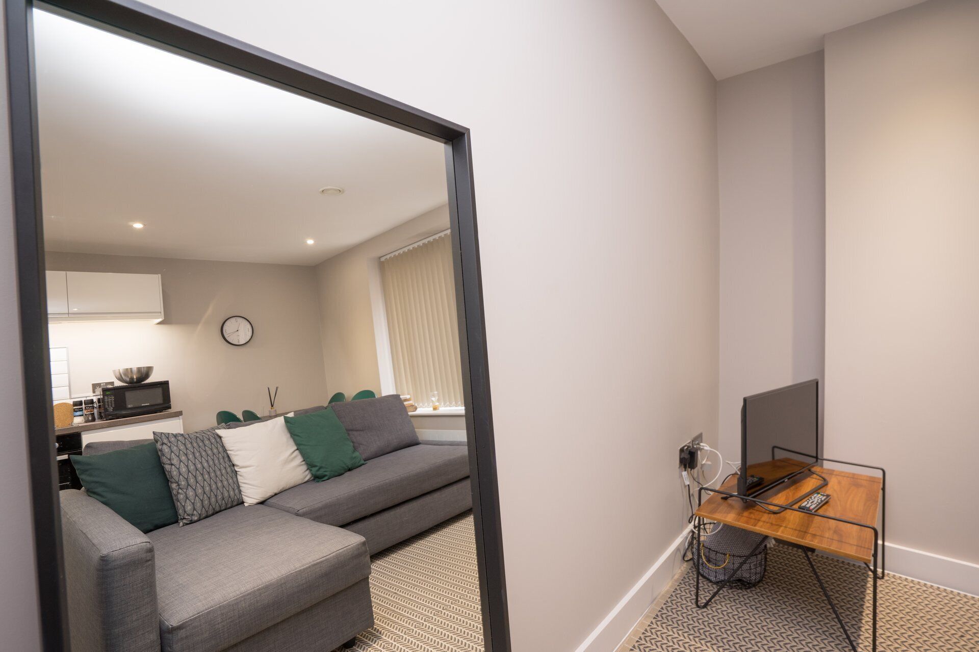 A modern living room with a grey sofa, a wooden side table, and a mirror reflecting the room.
