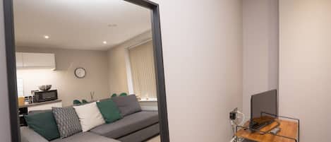 A modern living room with a grey sofa, a wooden side table, and a mirror reflecting the room.
