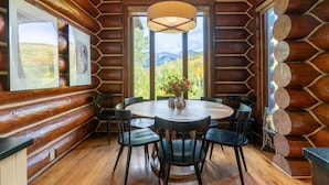 Une salle à manger aménagée dans une cabane en rondins, avec une table ronde, des chaises et une grande fenêtre donnant sur un paysage de montagnes et les couleurs automnales des arbres.