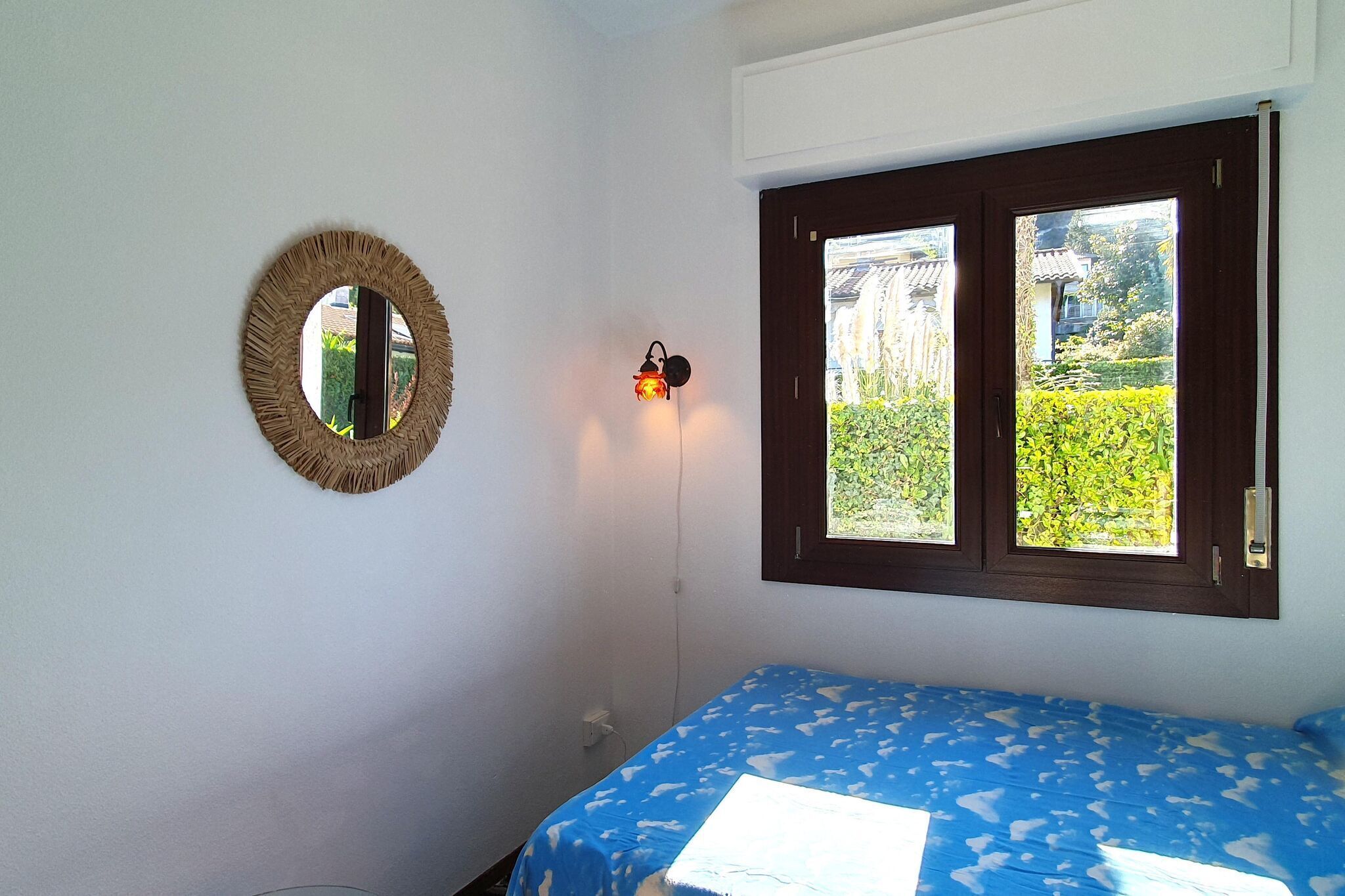 A room with a bed, a window with a view of greenery, a wall-mounted mirror, and a wall light.