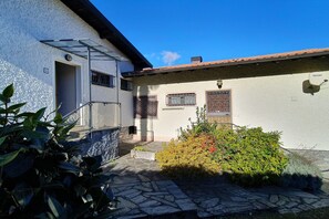 Ein Gebäude mit einer Steintreppe, die zu einer Tür führt, ein kleiner Garten mit verschiedenen Pflanzen und einem wolkenlosen, blauen Himmel.