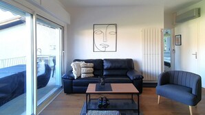 Un salon moderne avec un canapé en cuir noir, une petite table basse et un fauteuil bleu. Un grand tableau abstrait orne le mur, et une porte-fenêtre coulissante donne accès à un espace extérieur.