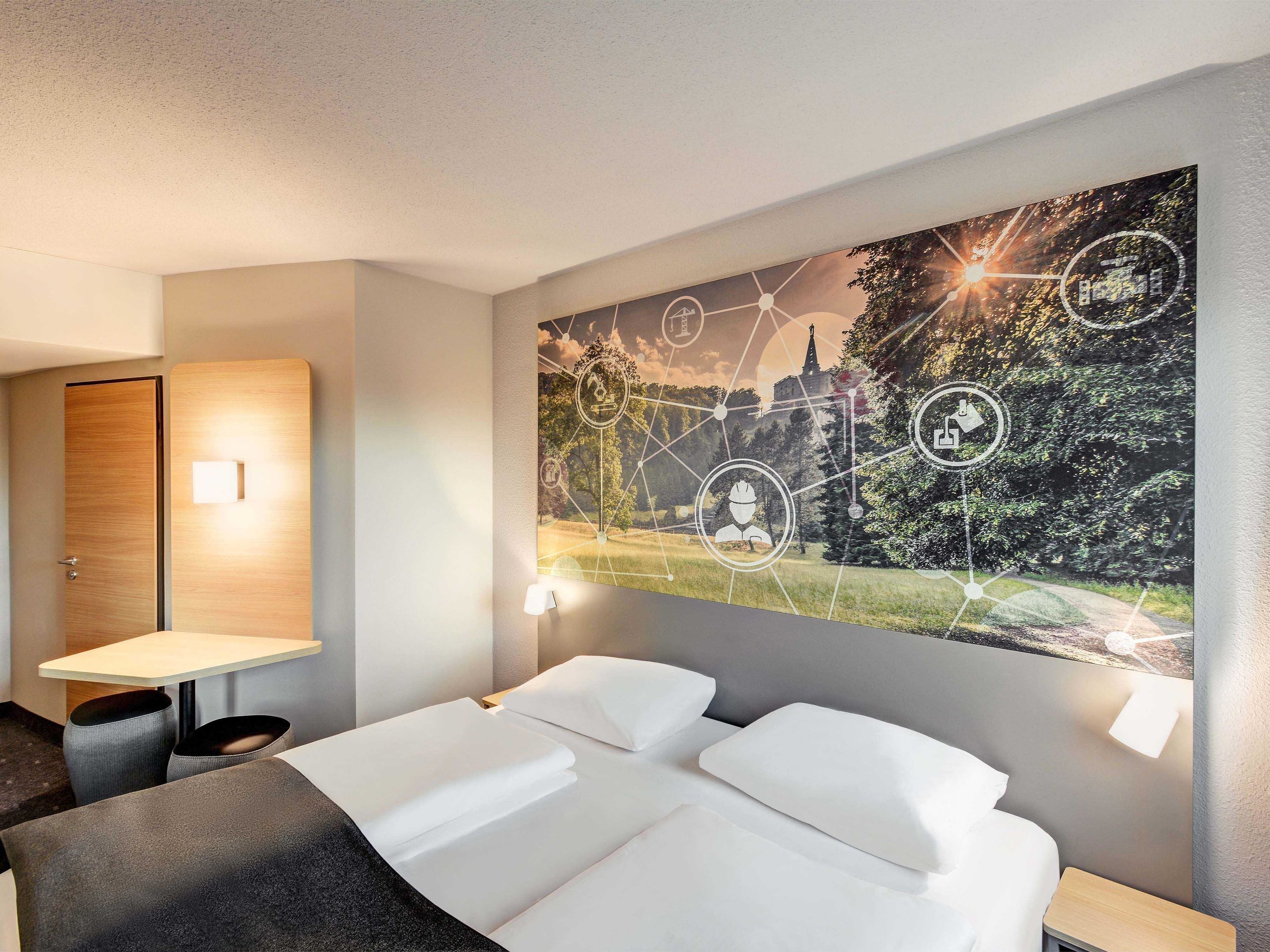 Une chambre d’hôtel avec une grande fresque murale, un lit avec du linge de maison blanc, un bureau avec une chaise et une porte en bois.