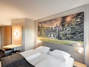 Hotelkamer met een groot wanddecor, een bed met witte lakens, een bureau met stoel en een houten deur.