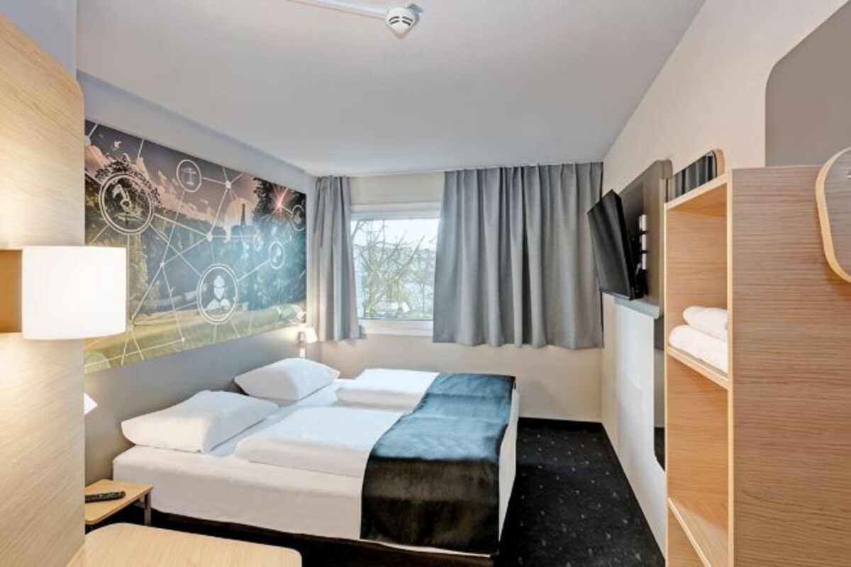 Une chambre d’hôtel comprenant un lit, un bureau, une télévision, une fenêtre avec des rideaux et un mur orné d’un motif géométrique.