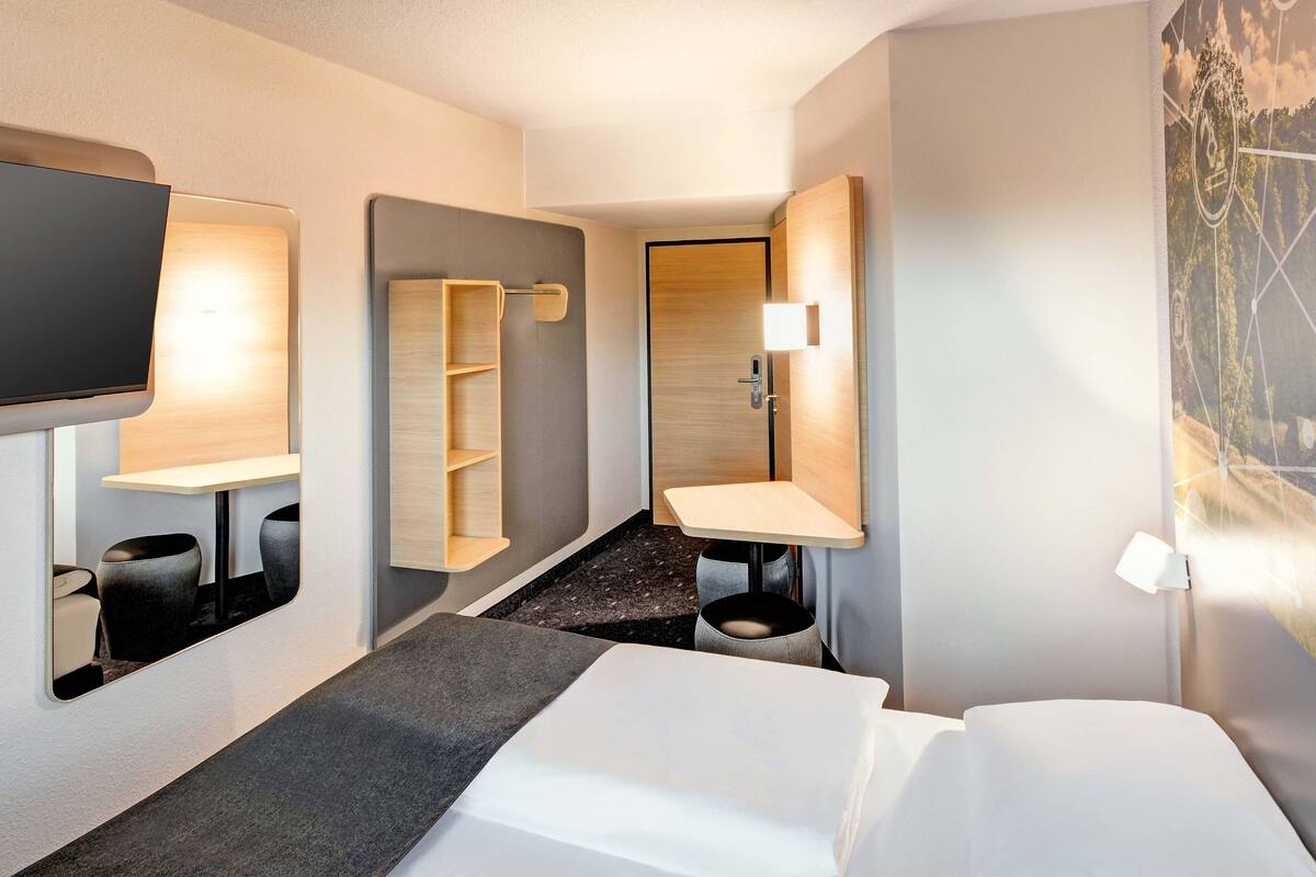 Une chambre d’hôtel moderne équipée d’un lit, d’une télévision, d’un bureau avec une chaise, d’un miroir et d’une salle de bain avec un lavabo et des toilettes.