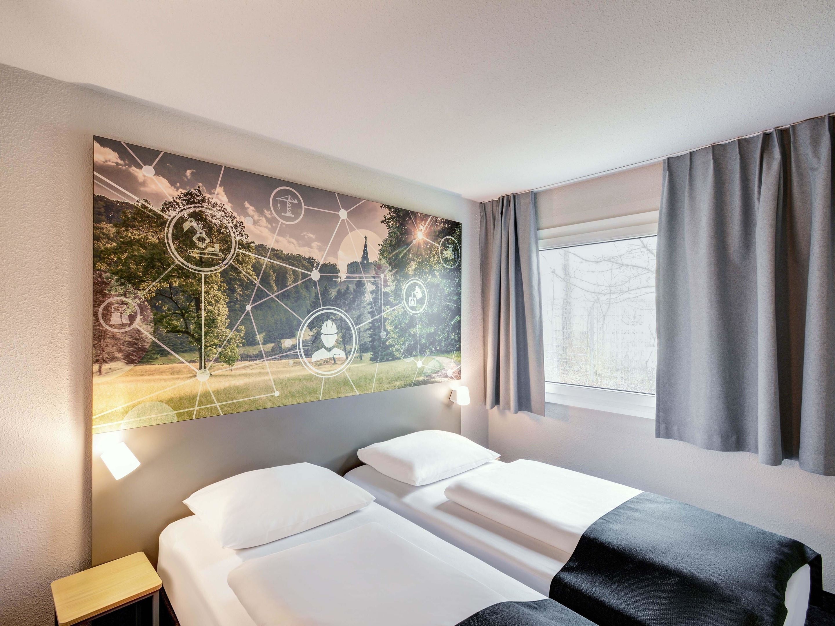 Ein Hotelzimmer mit zwei Betten, einem Nachttisch, einem an der Wand befestigten Kunstwerk, das eine Landschaft mit Symbolen zeigt, und einem Fenster mit Vorhängen.