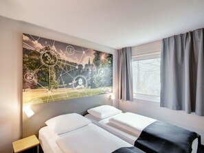 Ein Hotelzimmer mit zwei Betten, einem Nachttisch, einem an der Wand befestigten Kunstwerk, das eine Landschaft mit Symbolen zeigt, und einem Fenster mit Vorhängen.