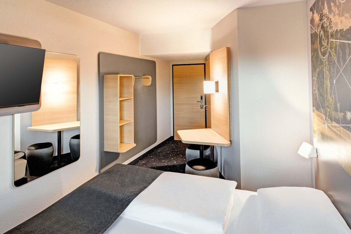 Une chambre d’hôtel moderne équipée d’un lit, d’une télévision, d’un bureau avec une chaise, d’un miroir et d’une salle de bain avec un lavabo et des toilettes.