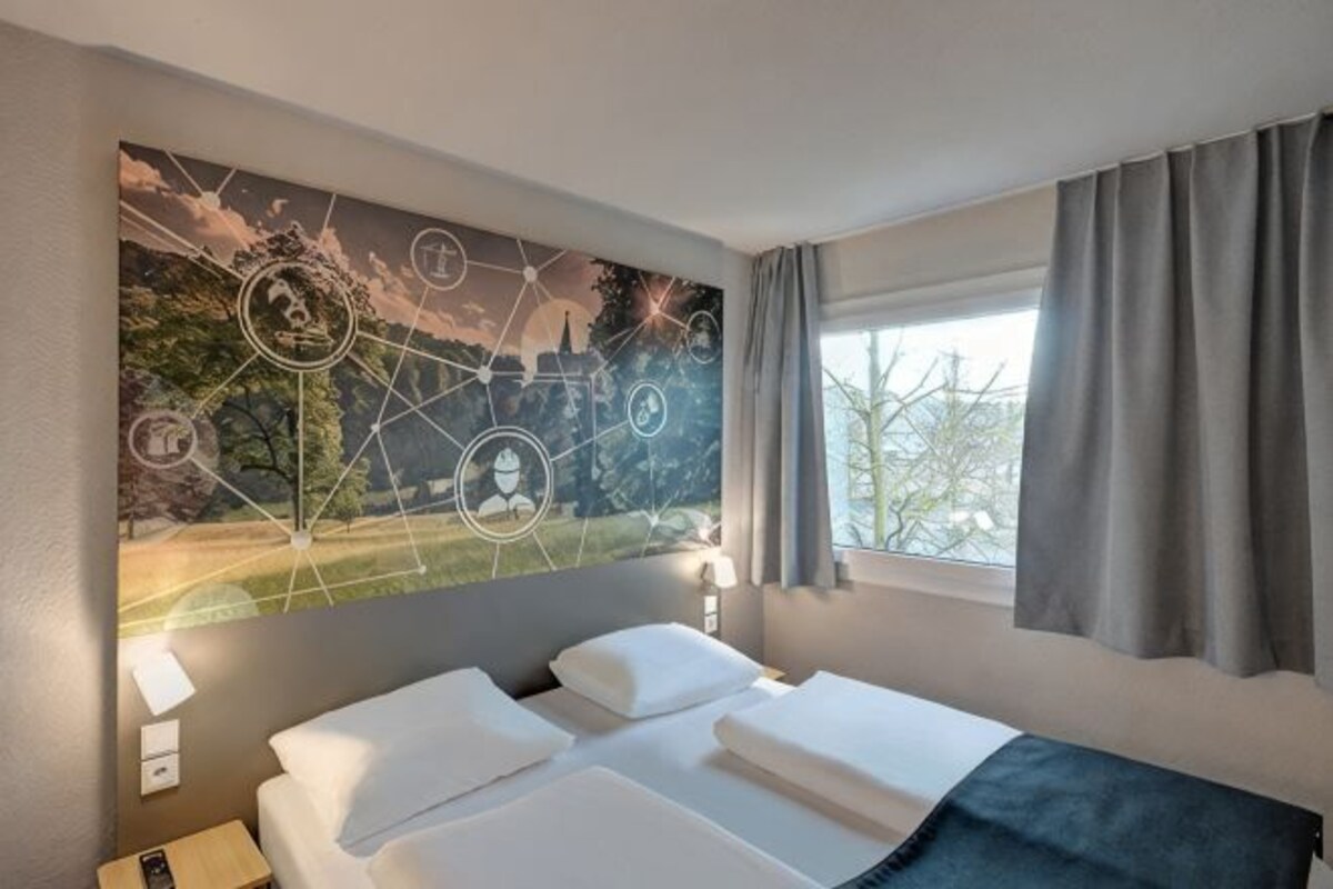 Une chambre d’hôtel avec un lit, une fenêtre avec des rideaux et un mur orné d’une œuvre d’art sur le thème de la nature.