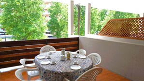 Mesa redonda com cadeiras brancas e toalha de mesa estampada, colocada numa varanda com vista para árvores e edifícios.