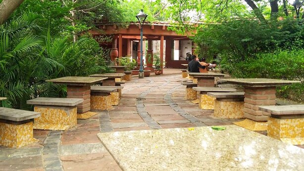 A garden pathway with stone benches and tables, surrounded by lush greenery and a red-roofed structure. - Roomshala 041 Hotel Sky Residency (New Delhi)