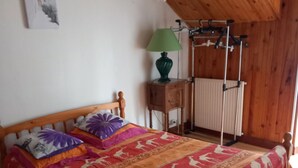 Un lit en bois surmonté d’une couvre-lit à motifs, une lampe verte et un radiateur dans une pièce aux murs en bois.