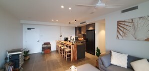 A modern living room with a sofa, a dining area with a wooden table and chairs, a kitchen with a refrigerator, and a ceiling fan. - New apartment, spectacular views day and night. Close to everything!! (Surfers Paradise)