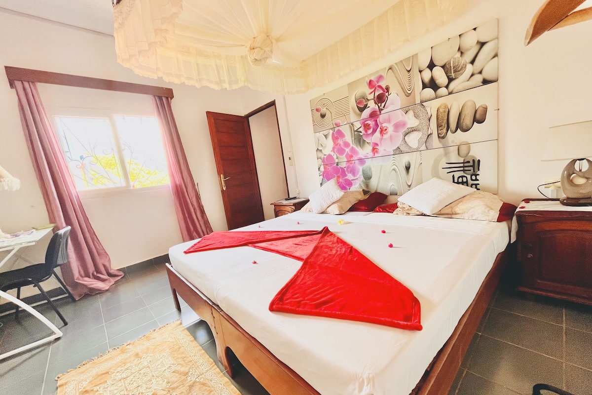 A bedroom with a bed, a desk, and a chair. There is a window with curtains, a door, and a wall with a decorative design.