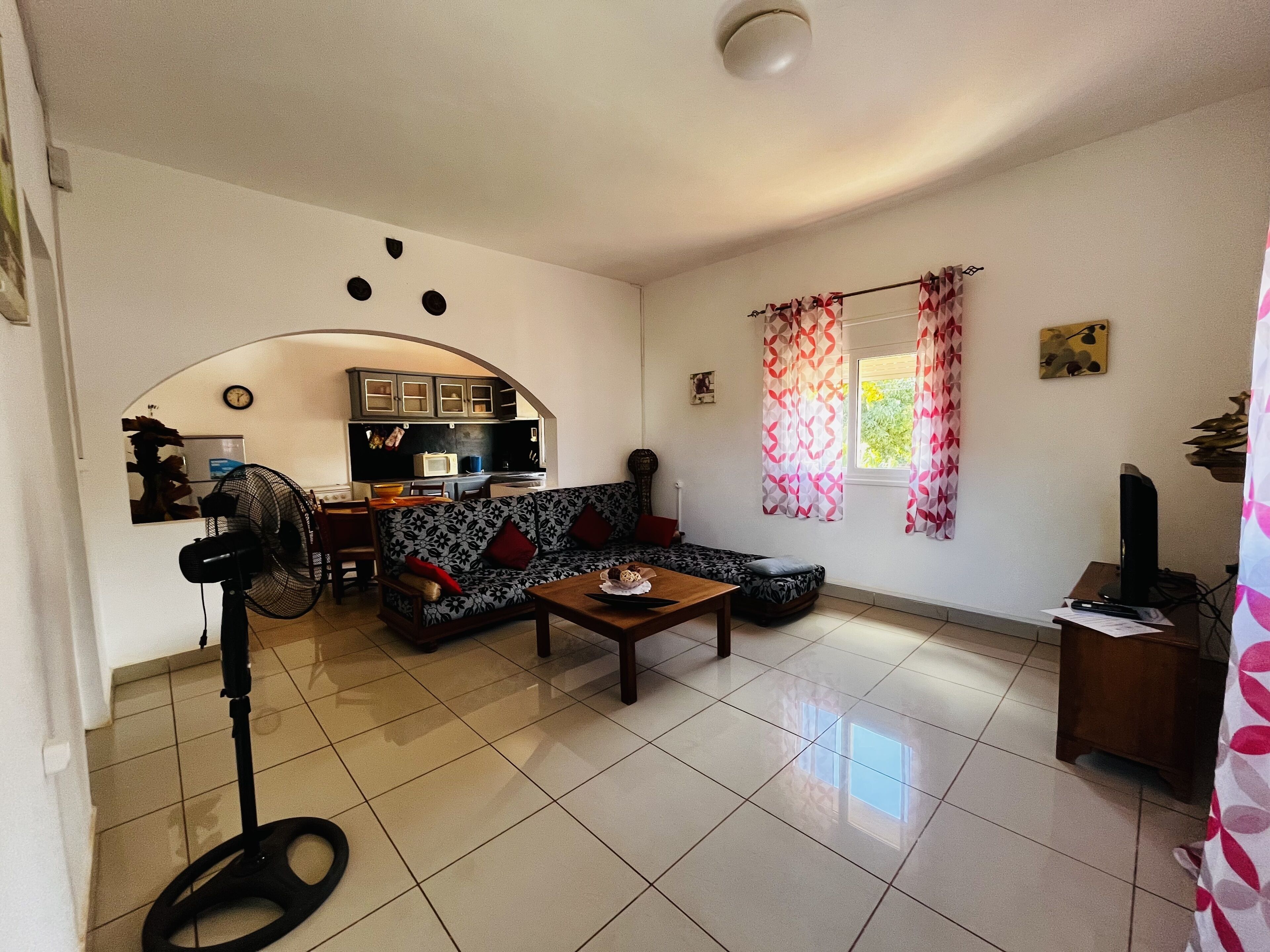 Een woonkamer met een bank, een salontafel, een ventilator en een keuken op de achtergrond.