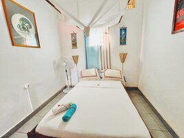 A hotel room with a bed, a standing fan, a curtain, and a painting on the wall.