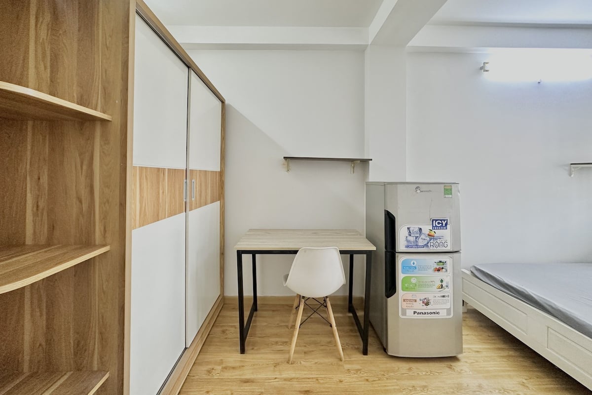 Een kleine, moderne kamer met een houten garderobe, een klein tafeltje met een stoel, een koelkast en een bed.