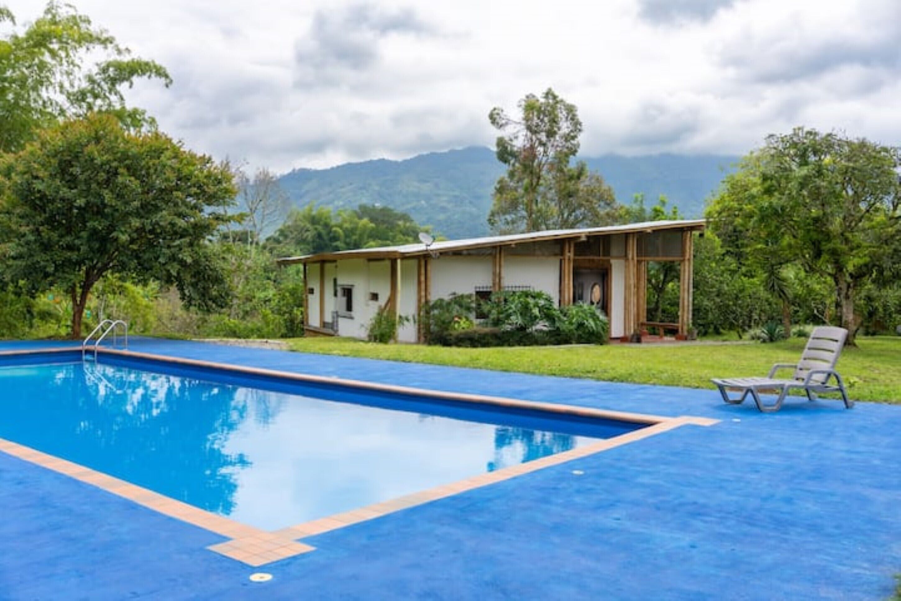 A swimming pool with a wooden deck and a lounge chair, surrounded by lush greenery and a mountain backdrop.
