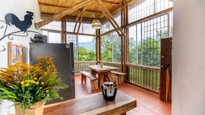 A wooden-framed balcony with a table and benches, a potted plant, and a rooster decoration on the wall.