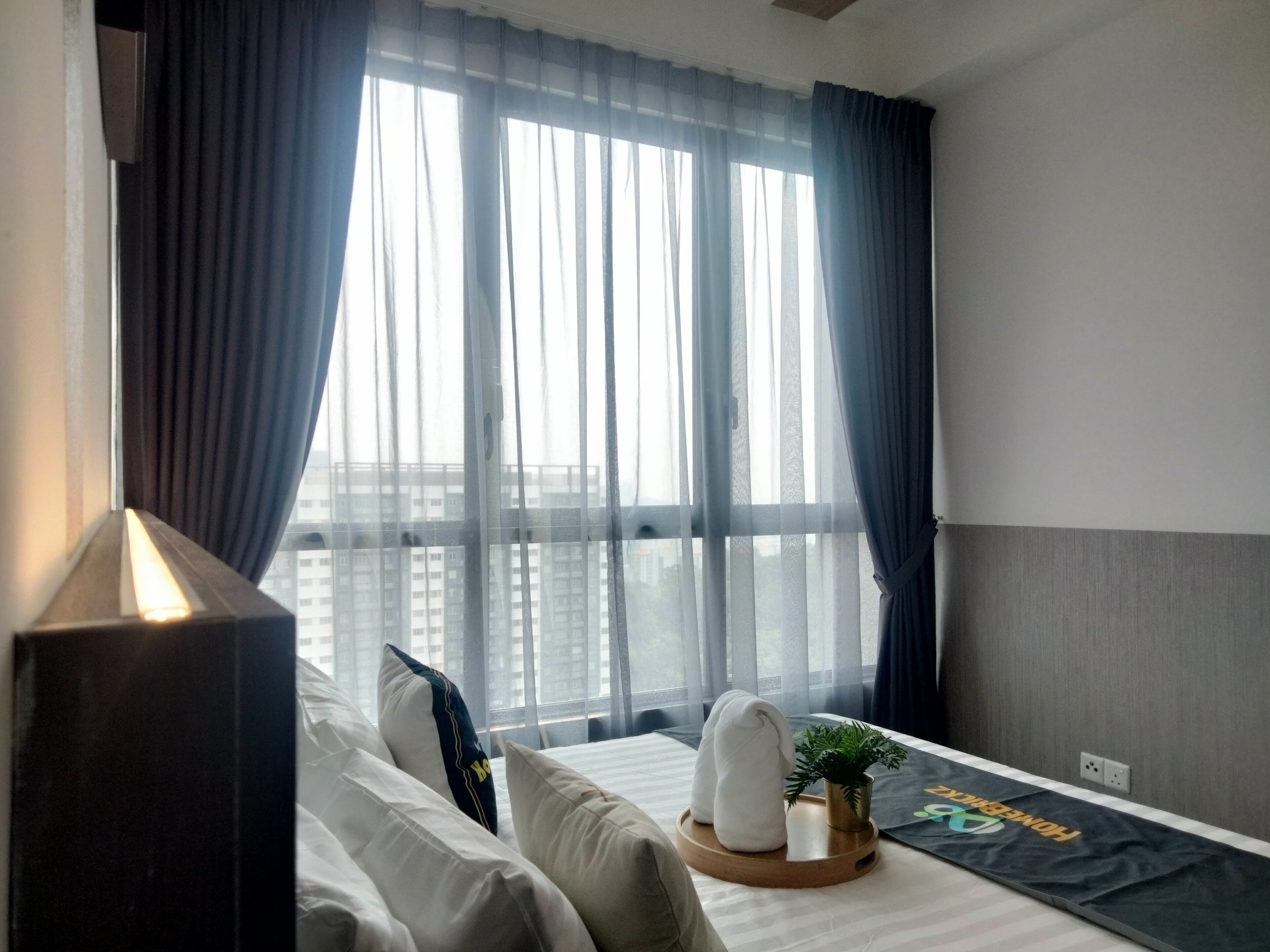 Habitació d'hotel amb llit, almoines, una taula de nit amb una planta i una finestra amb cortines transparents.