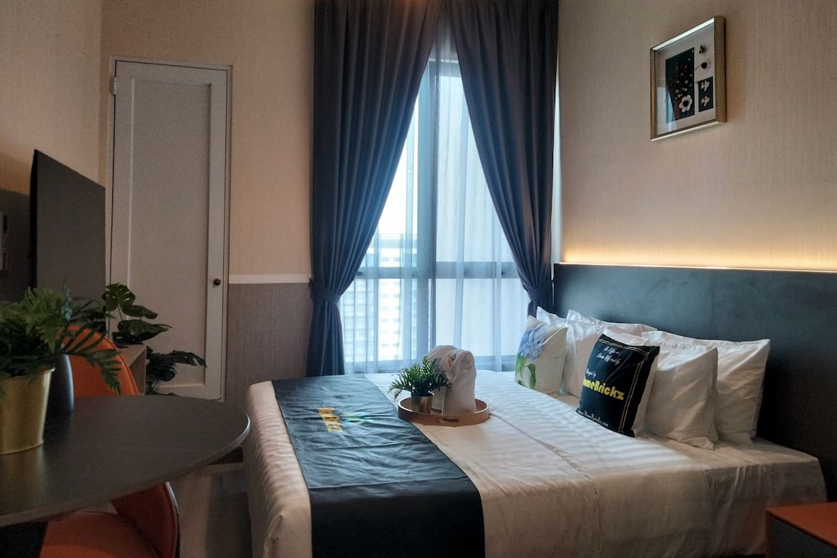 A hotel room with a bed, a desk, a chair, a potted plant, and a window with curtains.