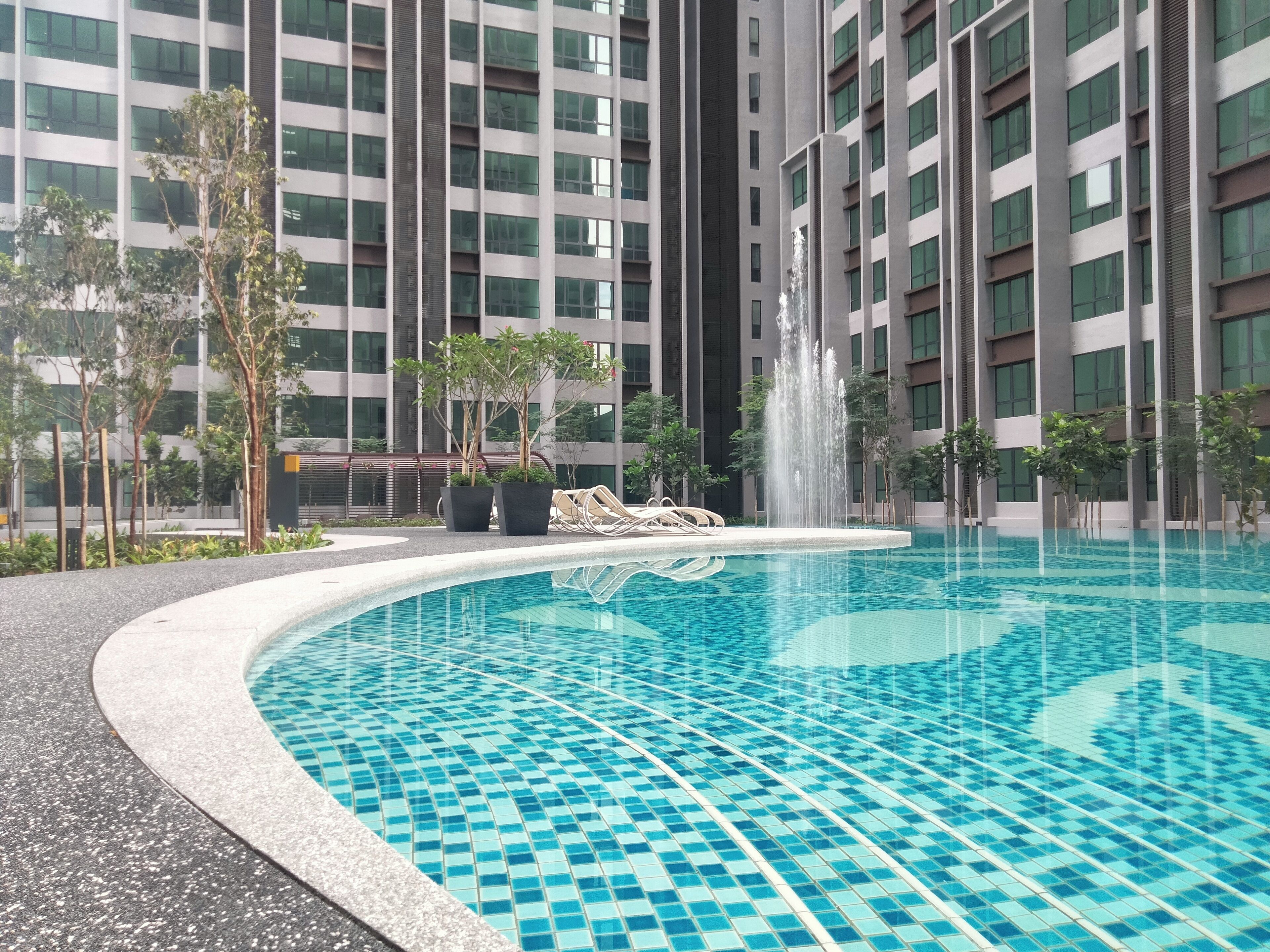 A swimming pool with a fountain, surrounded by trees and a modern building with multiple floors.