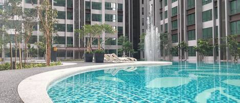 A swimming pool with a fountain, surrounded by trees and a modern building with multiple floors.