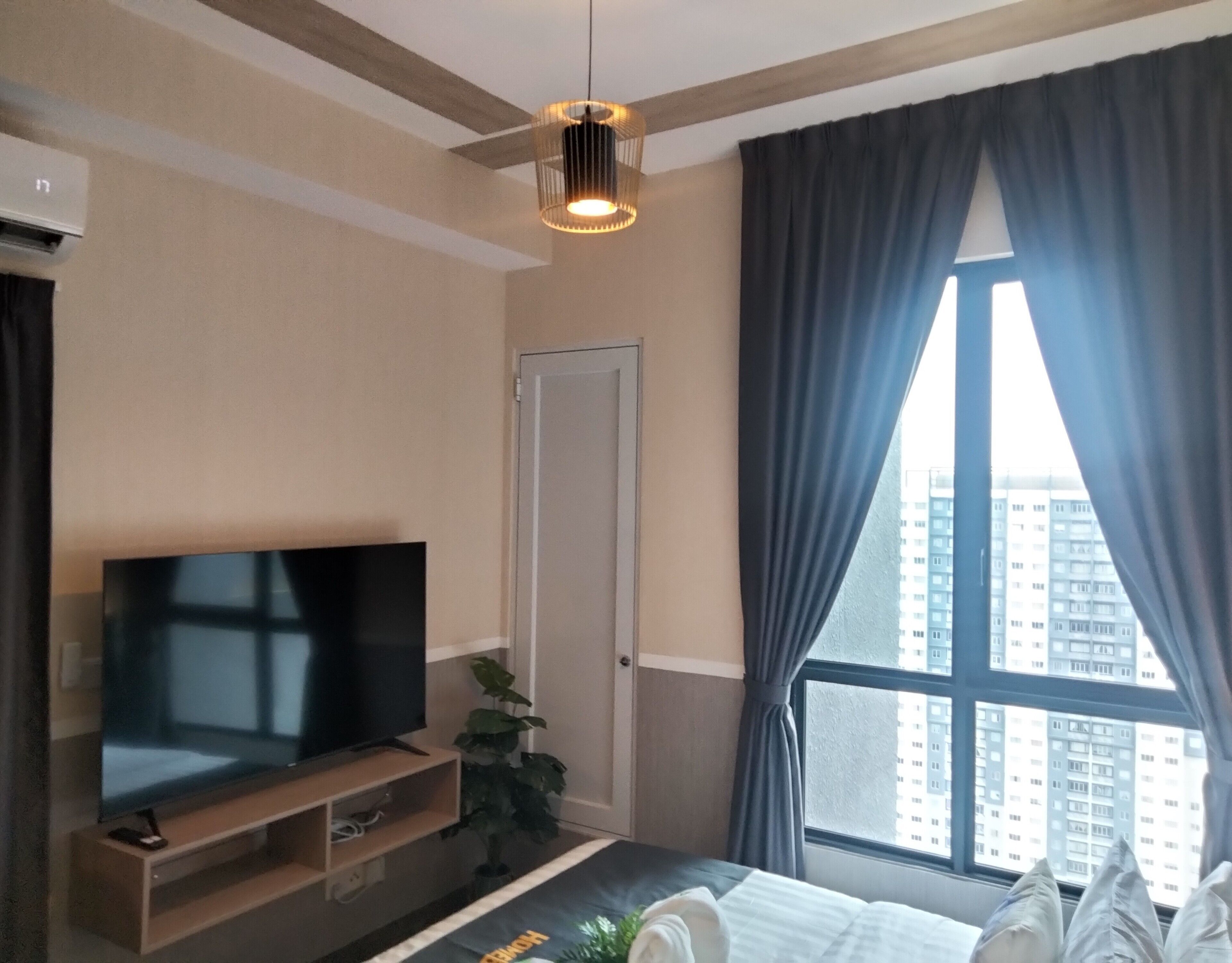 A modern hotel room with a flat-screen TV on a wooden stand, a potted plant, a bed with white bedding, and a large window with curtains.