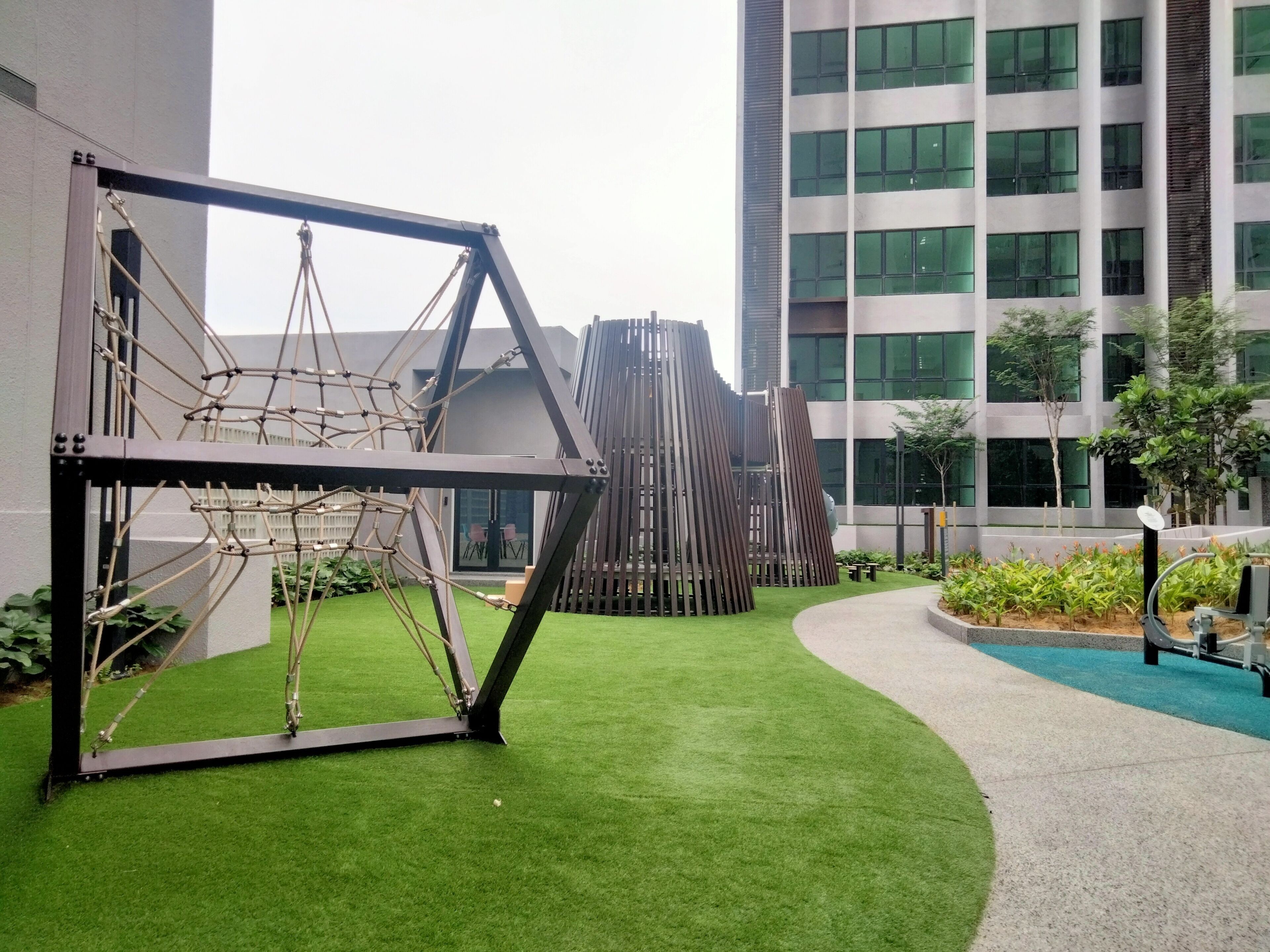 A playground with a rope bridge and climbing structure, surrounded by green artificial grass and a modern building in the background.