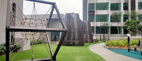 A playground with a rope bridge and climbing structure, surrounded by green artificial grass and a modern building in the background.