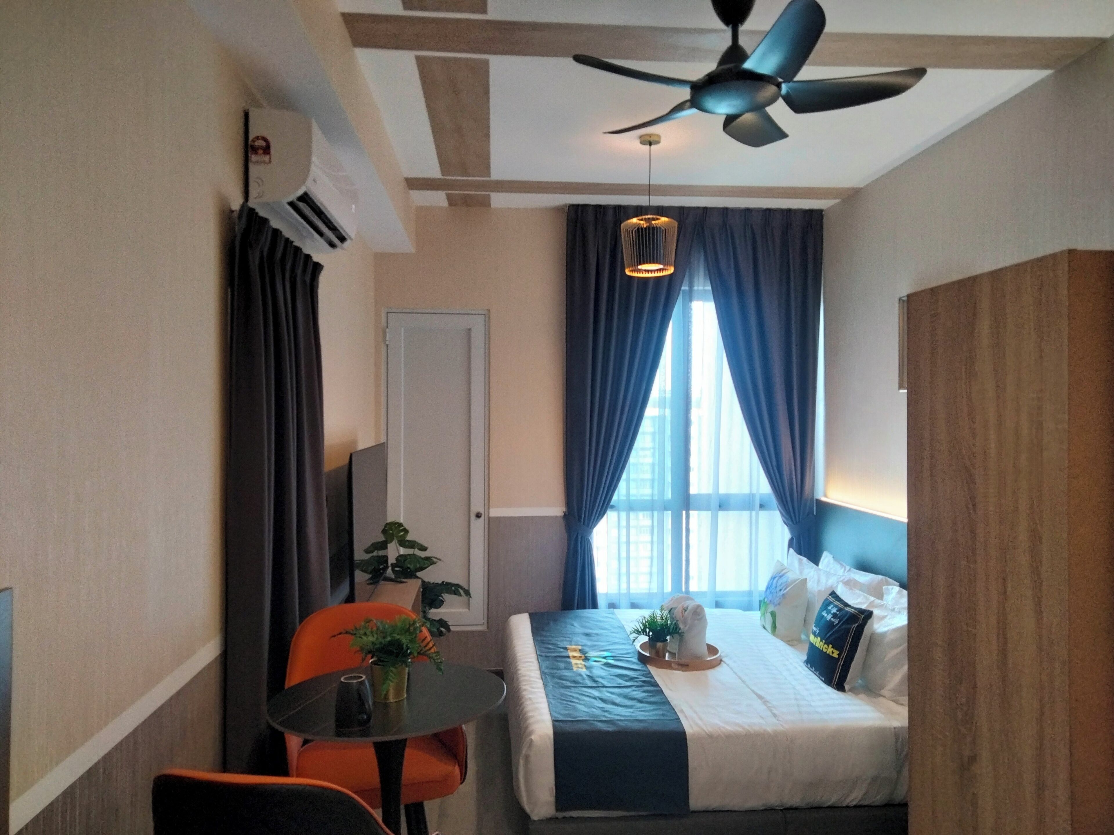 A hotel room with a bed, a desk, an orange chair, a ceiling fan, and a window with curtains.