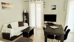 Ein modernes Wohnzimmer mit einer Couch, einem Couchtisch, einem Esstisch mit Stühlen, einem Fernseher, einem Bücherregal und einem Fenster mit Vorhängen.