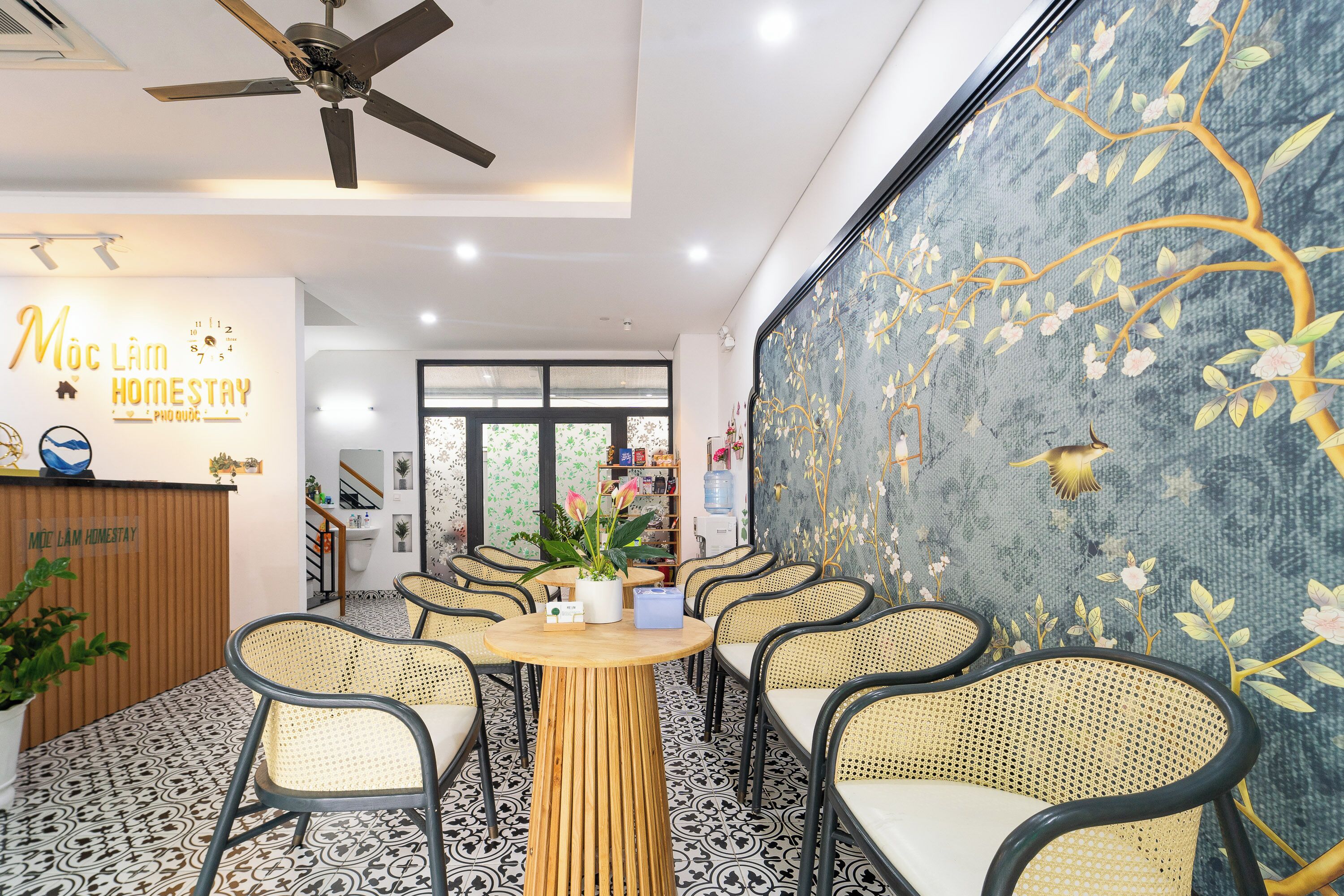 A cozy indoor seating area with wicker chairs and a central wooden table, surrounded by a patterned floor and a decorative wall mural.