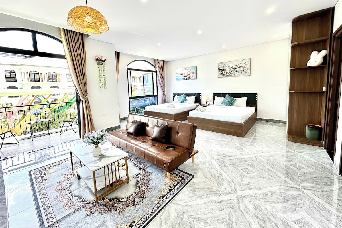 A hotel room with a balcony, two beds, a sofa, a coffee table, and a large window.