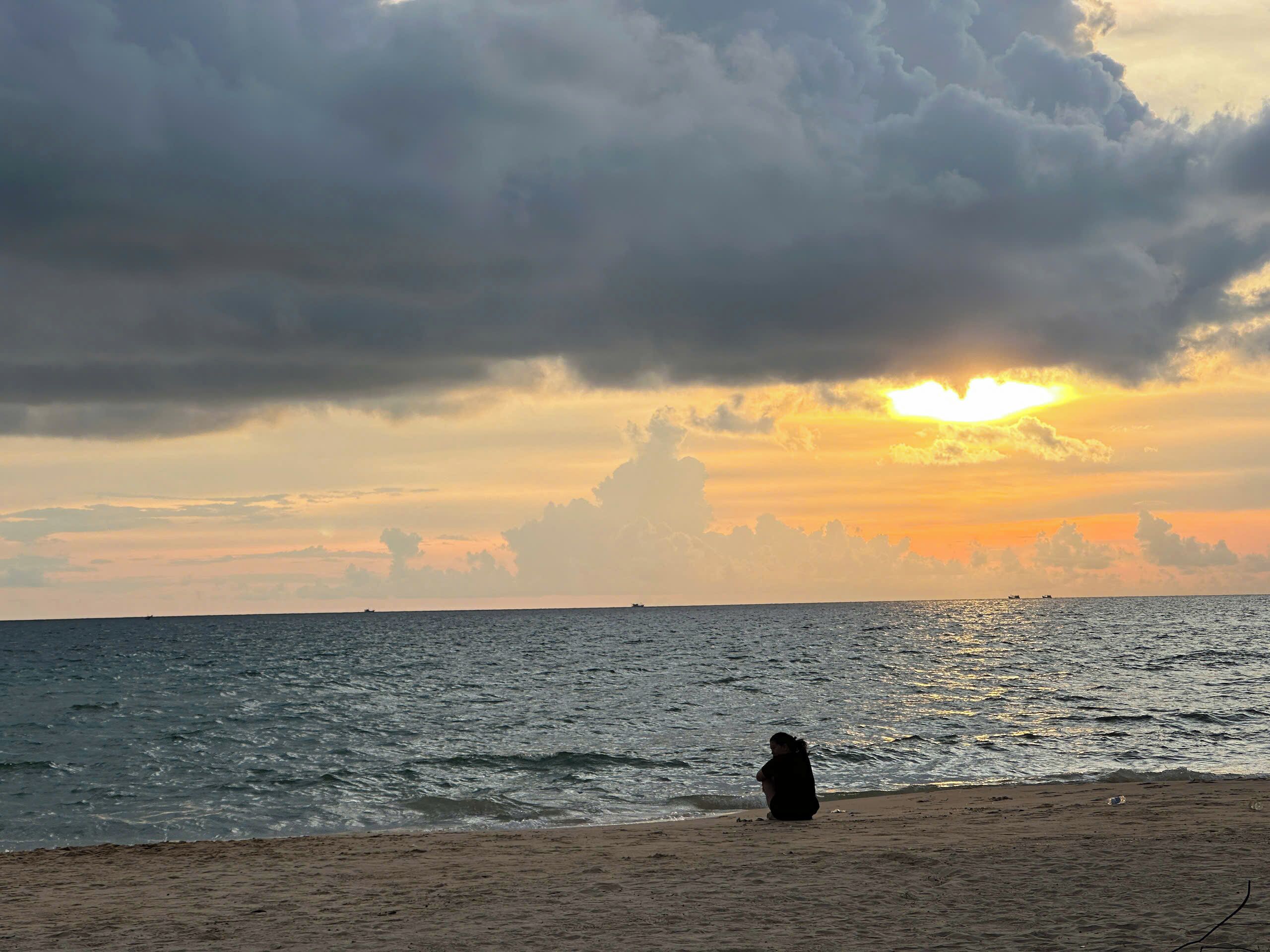 A beach with a person sitting on the sand, the ocean, and a sunset in the background.